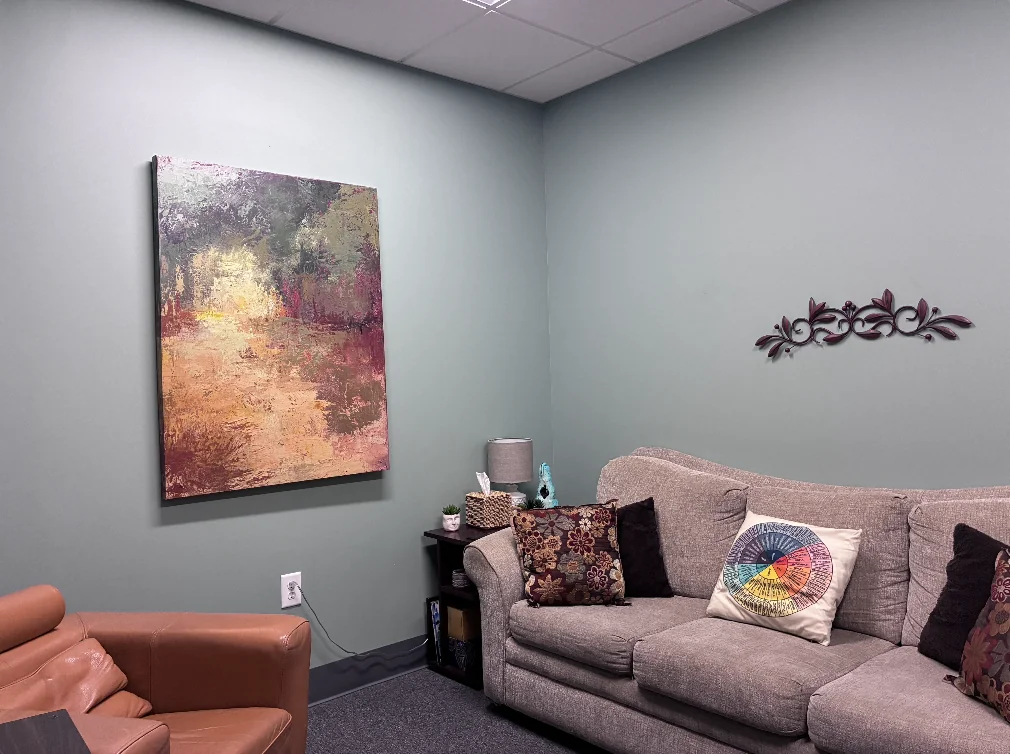 Office 1 with a gray sofa, a brown leather chair, patterned cushions, and artwork on muted green walls.