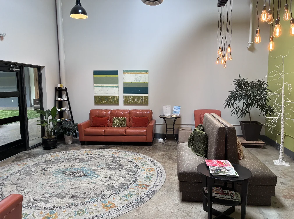 Waiting room with red and gray seating, plants, pendant lights, and a round rug.